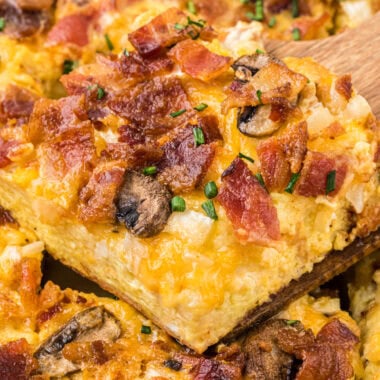 This Easy Bacon Breakfast Casserole has a creamy, cheesy custard base and a topping filled with crisp, smoky bacon, mushrooms, and onion. Easy to assemble, it can be prepared just before serving or made ahead the night before. This casserole is a favorite for weekend brunches, breakfast for dinner, or holiday breakfasts. Here the casserole is shown with a slice on a wooden spatula in the center of the image above the casserole.