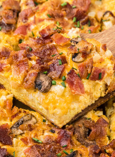 This Easy Bacon Breakfast Casserole has a creamy, cheesy custard base and a topping filled with crisp, smoky bacon, mushrooms, and onion. Easy to assemble, it can be prepared just before serving or made ahead the night before. This casserole is a favorite for weekend brunches, breakfast for dinner, or holiday breakfasts. Here the casserole is shown with a slice on a wooden spatula in the center of the image above the casserole.
