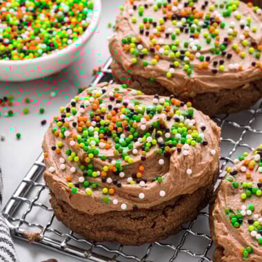 These Chocolate Frosted Cookies have a rich chocolate chocolate chip cookie base and the most creamy, decadent chocolate frosting. We love decorating them with sprinkles depending on the season. These cookies are a chocolate lover's dream and are perfect for the holidays, potlucks, or anytime chocolate cravings. Here the cookies are placed on a small cooling rack placed at a diagonal with a small bowl of sprinkles and a cookie in the background.