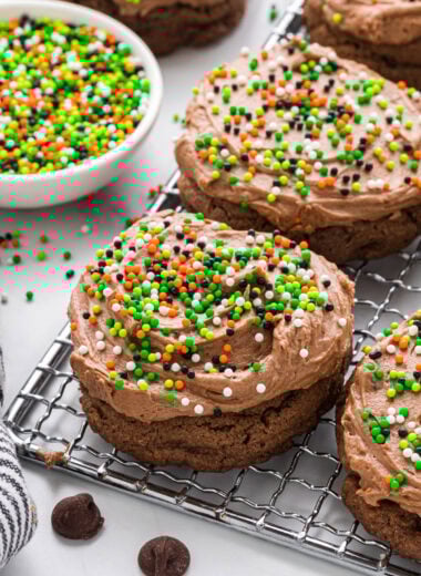 These Chocolate Frosted Cookies have a rich chocolate chocolate chip cookie base and the most creamy, decadent chocolate frosting. We love decorating them with sprinkles depending on the season. These cookies are a chocolate lover's dream and are perfect for the holidays, potlucks, or anytime chocolate cravings. Here the cookies are placed on a small cooling rack placed at a diagonal with a small bowl of sprinkles and a cookie in the background.