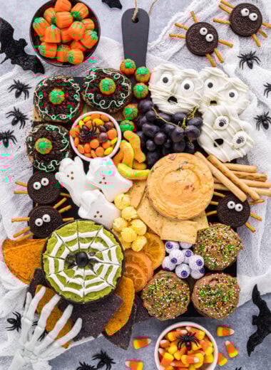 This Halloween Themed Charcuterie Board is as much fun to see as it is to eat. This kid-friendly snack board is filled with sweet and savory Halloween treats, and has a playful mix of spooky, silly, and sweet. The recipes for the cookies, mummy pretzels, and spider web dip are in the blog post as well as lots of tips for how to create a charcuterie board. This board is easy to customize and is a favorite at any Halloween table, party, or gathering. This overhead shot of the board shows Pumpkin Patch Cookies, Chocolate Frosted Cookies, Mummy Pretzels, and a Spider Web dip in each corner of the board. The board is filled with Oreo Spider Cookies, a pimento cheese dip, grapes, cheddar popcorn, dried oranges, Halloween chips, pretzels, pita chips, gummy worms, marshmallow ghosts, candy pumpkins, and candy eyeballs.