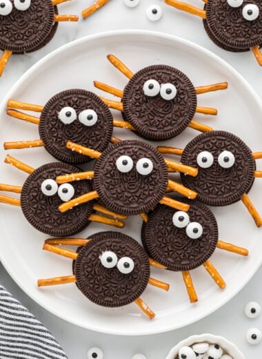 These Oreo Cookie Spiders are an easy, kid-friendly Halloween treat. Made with Oreo cookies, pretzel sticks for legs, and candy eyes, these cute spiders have their own little personality. Perfect for a Halloween party, a spooky school lunch snack, or a fun activity to do with kids, these Halloween spiders are as fun to make as they are to eat. This is an overhead image with the cookies on a plate in the center of the image, with additional spider cookies, pretzels and candy eyeballs around the plate.