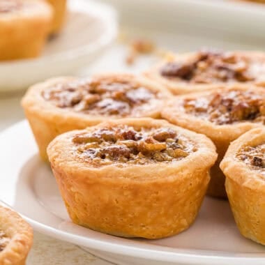These Pecan Tassies with Honey have a buttery cream cheese crust and a rich, gooey pecan filling. They are bite-sized treats that taste like mini pecan pies and are a perfect addition to any holiday dessert platter. In this image, the tassies are arranged on a plate in the center of the image with a plate and platter of tassies in the background and some finely chopped pecans in the foreground and background.