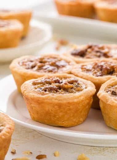 These Pecan Tassies with Honey have a buttery cream cheese crust and a rich, gooey pecan filling. They are bite-sized treats that taste like mini pecan pies and are a perfect addition to any holiday dessert platter. In this image, the tassies are arranged on a plate in the center of the image with a plate and platter of tassies in the background and some finely chopped pecans in the foreground and background.