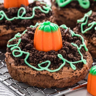 These Pumpkin Patch Cookies are a fun fall favorite. Made with our Chocolate Frosted Cookies, Oreo cookie crumbs, candy pumpkins, and white chocolate vines, they are cute little mini pumpkin patch treats. Kids love both making and eating them, and they are fun to share with friends, classmates, or at Halloween and fall parties. Here the cookies are placed on a cooling rack with additional candy pumpkins next to and behind the cookies.