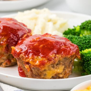 This Cheesy Mini Meatloaf is a kid-friendly weeknight dinner that cooks so much faster than a larger meatloaf. Each mini meatloaf is juicy, packed with flavor, and has everything you love about a traditional meatloaf in muffin-sized form. The cheesy goodness in each bite, combined with the tender meat and ketchup-based sauce, makes these a year-round family favorite. Here, there are two cheesy mini meatloaves plated in the center of the image with mashed potatoes and broccoli, with an additional plate in the background as well as additional sauce for serving.