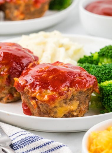 This Cheesy Mini Meatloaf is a kid-friendly weeknight dinner that cooks so much faster than a larger meatloaf. Each mini meatloaf is juicy, packed with flavor, and has everything you love about a traditional meatloaf in muffin-sized form. The cheesy goodness in each bite, combined with the tender meat and ketchup-based sauce, makes these a year-round family favorite. Here, there are two cheesy mini meatloaves plated in the center of the image with mashed potatoes and broccoli, with an additional plate in the background as well as additional sauce for serving.