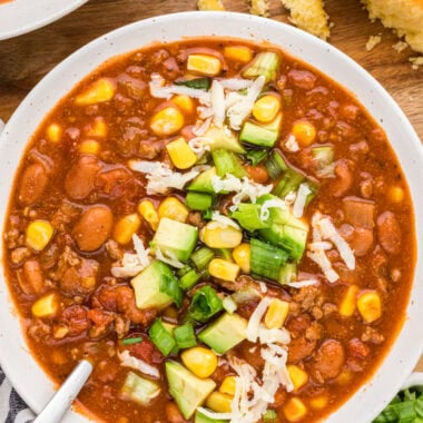 Just last night, I won first place in a chili contest with this Chili Recipe with Corn. We love this chili. It is the perfect one-pot meal. It makes a great cozy dinner on a busy weeknight, or easily feeds a crowd for game nights, Halloween, or other fall and winter gatherings. Made with ground beef, chili beans, pinto beans, green chiles, broth, tomatoes, tomato sauce, seasonings, and corn, we love how the slight sweetness of the corn balances with the flavorful seasoned chili. This image shows an overhead of the chili served in a bowl with toppings including corn, avocao, green onions and grated cheese. Cornbread is shown above the bowl of chili.