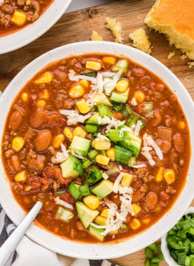 Just last night, I won first place in a chili contest with this Chili Recipe with Corn. We love this chili. It is the perfect one-pot meal. It makes a great cozy dinner on a busy weeknight, or easily feeds a crowd for game nights, Halloween, or other fall and winter gatherings. Made with ground beef, chili beans, pinto beans, green chiles, broth, tomatoes, tomato sauce, seasonings, and corn, we love how the slight sweetness of the corn balances with the flavorful seasoned chili. This image shows an overhead of the chili served in a bowl with toppings including corn, avocao, green onions and grated cheese. Cornbread is shown above the bowl of chili.