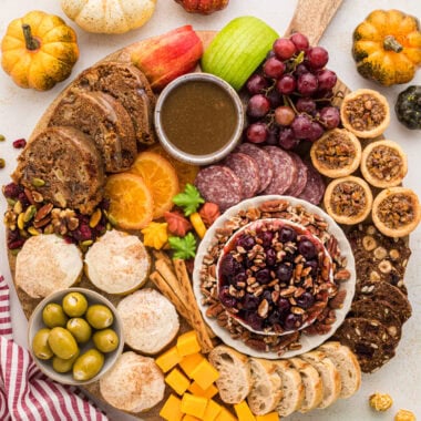 This Fall Themed Charcuterie Board is the ultimate autumn centerpiece. Filled with sweet and savory fall desserts and snacks, it's perfect for Friendsgiving, Thanksgiving, or just a fun night at home. The recipes for the pecan tassies, pumpkin cream cheese cookies, apple caramel cake, caramel sauce, and cherry pecan brie are in the blog post, as well as lots of tips for how to create a charcuterie board. This board is easy to customize using your favorite fall eats and treats. Here you see an overhead view of the round board filled with the baked goods mentioned as well as feta cheese stuffed olives, pretzels, sliced baguette, cranberry hazelnut crisps, trail mix, sliced dried oranges, sliced salami, cubed cheddar cheese, grapes, sliced apples and autumn leaf candies.