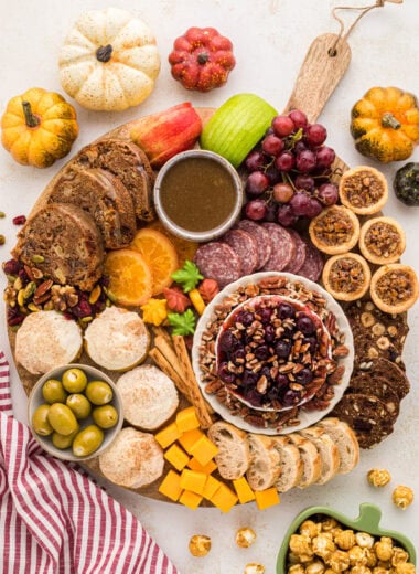 This Fall Themed Charcuterie Board is the ultimate autumn centerpiece. Filled with sweet and savory fall desserts and snacks, it's perfect for Friendsgiving, Thanksgiving, or just a fun night at home. The recipes for the pecan tassies, pumpkin cream cheese cookies, apple caramel cake, caramel sauce, and cherry pecan brie are in the blog post, as well as lots of tips for how to create a charcuterie board. This board is easy to customize using your favorite fall eats and treats. Here you see an overhead view of the round board filled with the baked goods mentioned as well as feta cheese stuffed olives, pretzels, sliced baguette, cranberry hazelnut crisps, trail mix, sliced dried oranges, sliced salami, cubed cheddar cheese, grapes, sliced apples and autumn leaf candies.