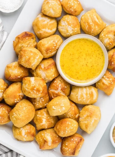 These Homemade Pretzel Bites are crispy on the outside and pillowy soft on the inside. Made with a few pantry ingredients, these bakery-quality pretzels are the best comfort snack and perfect for dipping and sharing. We love their crisp, salty crust and fluffy middle; they are impossible to stop eating and make a perfect after school, game day, fun day snack. Here the pretzel bites are shown served on a platter with honey mustard sauce, with other sauces and kosher salt around the platter in an overhead shot.