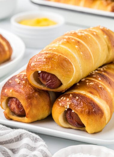 This Pretzel Dog Recipe is the ultimate mash-up of two comfort foods. The pillowy soft, salty dough wrapped around the savory, juicy hot dogs makes a kid-friendly, nostalgic treat. Perfect for fun family meals or game day snacks, both kids and adults love them. In this image, there are three hot dogs placed on a small platter, ready to serve with a few other pretzel dogs in the background as well as various mustards.