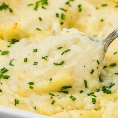 These Baked Mashed Potatoes with Cream Cheese are the ultimate comfort food and make the perfect side dish for holiday baking. These make-ahead potatoes can be made 1-2 days in advance and then baked just before serving. Made with cream cheese, butter in the potatoes and on top, and cream, they are ultimately creamy and buttery, with a light golden top. Here a spoon is scooping the potatoes out of the pan in the center of the image