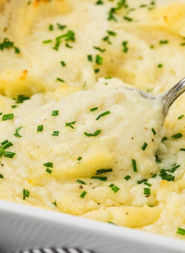 These Baked Mashed Potatoes with Cream Cheese are the ultimate comfort food and make the perfect side dish for holiday baking. These make-ahead potatoes can be made 1-2 days in advance and then baked just before serving. Made with cream cheese, butter in the potatoes and on top, and cream, they are ultimately creamy and buttery, with a light golden top. Here a spoon is scooping the potatoes out of the pan in the center of the image