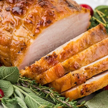 This Brined Boneless Turkey Breast with Maple Glaze is incredibly tender and flavorful. The overnight brine, seasoning, and maple butter glaze when roasting, gives this boneless turkey breast an incredible depth of flavor and just the right balance of slightly sweet and savory. This is an easy, elegant dish for dinner or for a small group for Thanksgiving if you don't want to make a large turkey. This is an overhead shot of the boneless turkey breast placed on a serving platter with sage, thyme, and cranberries. The breast has several slices cut so you can see the interior of the turkey as well.