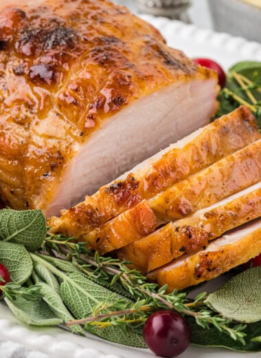 This Brined Boneless Turkey Breast with Maple Glaze is incredibly tender and flavorful. The overnight brine, seasoning, and maple butter glaze when roasting, gives this boneless turkey breast an incredible depth of flavor and just the right balance of slightly sweet and savory. This is an easy, elegant dish for dinner or for a small group for Thanksgiving if you don't want to make a large turkey. This is an overhead shot of the boneless turkey breast placed on a serving platter with sage, thyme, and cranberries. The breast has several slices cut so you can see the interior of the turkey as well.