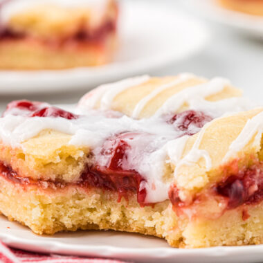 We make these Cherry Bars with Almond Glaze every holiday season. Made in a half sheet pan, they make a beautiful dessert for a larger crowd or make an easy Christmas gift. The tender crumb, cherry pie filling, and almond icing drizzle are a delicious combination, and they are easy to prepare. In this image, a cherry bar is plated in the center of the image with a bite taken, showing the inside tender crumb, cherry pie filling, and almond drizzle up close. Plated bars are in the background.