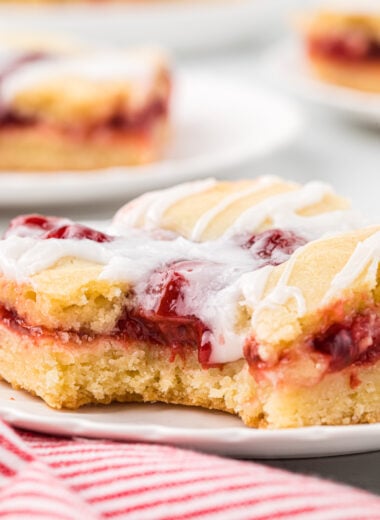We make these Cherry Bars with Almond Glaze every holiday season. Made in a half sheet pan, they make a beautiful dessert for a larger crowd or make an easy Christmas gift. The tender crumb, cherry pie filling, and almond icing drizzle are a delicious combination, and they are easy to prepare. In this image, a cherry bar is plated in the center of the image with a bite taken, showing the inside tender crumb, cherry pie filling, and almond drizzle up close. Plated bars are in the background.