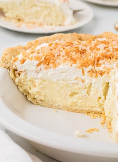 This Coconut Cream Pie with Coconut Milk, made with part coconut milk and half-and half, is my husband's favorite pie. We have made it for countless birthdays and holidays, and truthfully, it is always a favorite for guests and family. It has a dreamy, creamy custard with coconut flavor that shines through from the coconut milk, sweetened coconut flakes in the custard, and toasted coconut flakes sprinkled on top. Here the pie is in the pan in the center of the image, and has been sliced into to see the creamy filling of the pie. Slices of the pie are in the background.