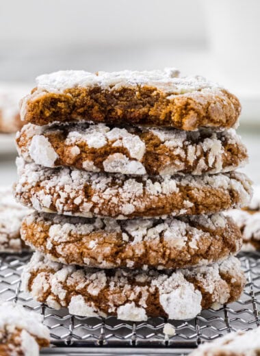 These Ginger Crinkle Cookies taste like Christmas. They have a crisp exterior, a powdered sugar shell, and a soft center with cozy gingerbread spices in every bite. The snowy, powdered sugar-cracked tops makes these cookies extra festive. We make these cookies every year, and they are loved by kids and adults alike. Here the cookies are stacked in the center of the image with a bite taken out of the top cookie to see the soft interior of the cookie