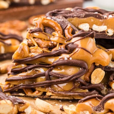 This Caramel Pretzel Chocolate Bark is the ultimate sweet-and-salty treat. The combination of the creamy chocolate base, crunchy and salty pretzels and peanuts, and a rich buttery caramel topping finished with a sprinkle of salt is an irresistible treat. Easy to make with just a few ingredients, its a fun holiday treat, perfect for packing in tins or sharing on a plate. In this image, a piece of the Caramel Pretzel Bark is leaning in front of a stack of the bark in the center of the frame with pretzels, peanuts and other bark pieces in the foreground and background.