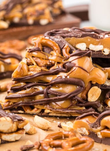 This Caramel Pretzel Chocolate Bark is the ultimate sweet-and-salty treat. The combination of the creamy chocolate base, crunchy and salty pretzels and peanuts, and a rich buttery caramel topping finished with a sprinkle of salt is an irresistible treat. Easy to make with just a few ingredients, its a fun holiday treat, perfect for packing in tins or sharing on a plate. In this image, a piece of the Caramel Pretzel Bark is leaning in front of a stack of the bark in the center of the frame with pretzels, peanuts and other bark pieces in the foreground and background.