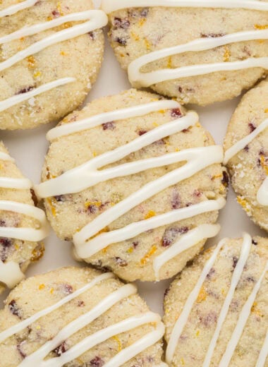 These Orange Cranberry Cookies with Icing are soft, citrusy, and sweet. Filled with tart dried cranberries, crispy chopped pecans, and bright citrusy orange zest, then rolled in orange sugar and drizzled with orange icing, they are a beautiful and delicious holiday treat. This image is an overhead shot of the iced cookies close together on a plate.