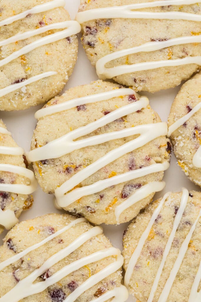 These Orange Cranberry Cookies with Icing are soft, citrusy, and sweet. Filled with tart dried cranberries, crispy chopped pecans, and bright citrusy orange zest, then rolled in orange sugar and drizzled with orange icing, they are a beautiful and delicious holiday treat. This image is an overhead shot of the iced cookies close together on a plate.