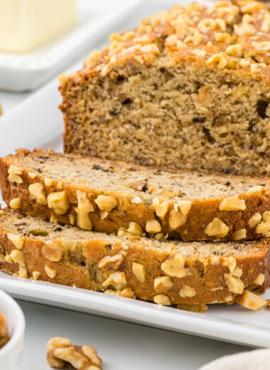 This Buttermilk Banana Nut Bread has the perfect combination of a tender, rich crumb from the buttermilk, sweetness from the ripe bananas, and a delicate crunch from the walnuts in the bread, and roasted on top. It's comfort in every bite. The hardest thing about this recipe is waiting for it to cool to eat it! A huge family favorite. This image shows the bread on a platter, with two slices cut in front of the bread. Butter and additional walnuts are in the foreground and background.