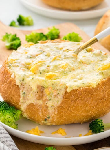 This Creamy Broccoli and Cheddar Soup is pure comfort in a bowl. The creamy, cheesy soup base with the tender broccoli is the perfect combination. We love this served with bread or in a bread bowl. In this image, the soup is served in a bread bowl in the center of the image, with a spoon in the bowl in the center of the image.