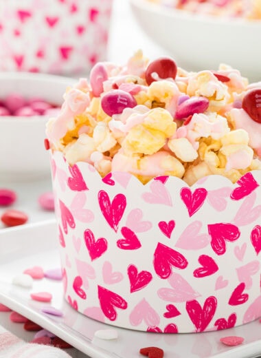 This White Chocolate Popcorn Valentine's mix is a cheerful, sweet, and crunchy treat. Drizzled in white chocolate and mixed with M&Ms and sprinkles, the popcorn is highly addictive, easy to make, and perfect for Valentine's parties, movie nights, and gifting. Here the popcorn mix is being served in a Valentine's Day paper cup with hearts on a tray in the center of the image, with additional m&Ms, popcorn cups, sprinkles, and the bowl of popcorn in the background.
