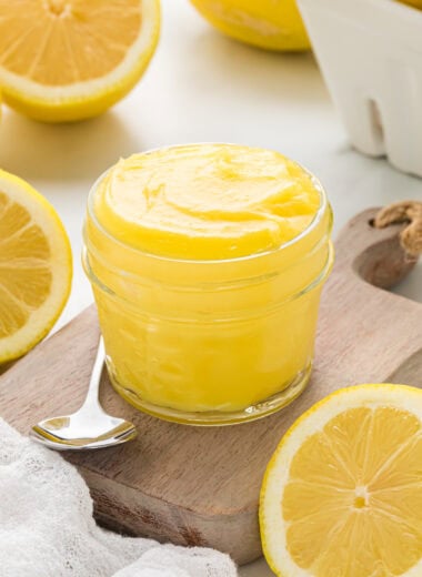 This lemon curd is the Best Lemon Curd Recipe with its rich, creamy custard and its perfect balance of sweet and tart. We love using this as a breakfast spread, as a filling for cookies and tarts, and as a topping with berries and whipped cream. It is absolutely irresistible and is the perfect, cheerful spring accompaniment to a variety of dishes and desserts. In this image, the lemon curd is in a small jar sitting on a small wooden board with a spoon ready to serve. Lemons are in the background and foreground.