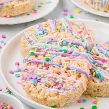 These Easter Egg Rice Krispies Treats get a boost of flavor from browned butter and vanilla, and are decorated with tinted white chocolate and sprinkles. They are an easy, no-bake, fun, and festive Easter treat that kids love making and eating. Here the Easter Egg Rice Krispies Treats are plated ready to eat. The center main image focuses on a treat stacked on two others on a small serving plate with two other smaller plates with treats in the background. Sprinkles and marshmallows are in the foreground and background.