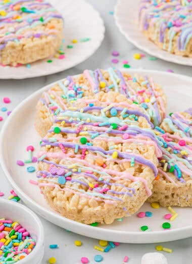 These Easter Egg Rice Krispies Treats get a boost of flavor from browned butter and vanilla, and are decorated with tinted white chocolate and sprinkles. They are an easy, no-bake, fun, and festive Easter treat that kids love making and eating. Here the Easter Egg Rice Krispies Treats are plated ready to eat. The center main image focuses on a treat stacked on two others on a small serving plate with two other smaller plates with treats in the background. Sprinkles and marshmallows are in the foreground and background.