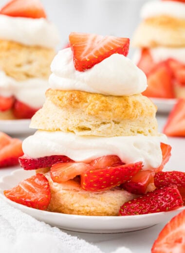 These Homemade Strawberry Shortcakes have a sweetened tender, buttery biscuit, a sweet strawberry filling, and a pillowy soft vanilla whipped cream topping. Perfect for spring and summer gatherings, this classic layered dessert is as stunning as it is delicious. Here the shortcakes are assembled and plated with a shortcake in the front center of the images and additional shortcakes and strawberries in the background
