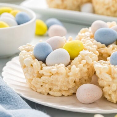 hese Easter Nests with Rice Krispies are an easy, no-bake, fun, and festive Easter treat that kids love making and eating. They get a boost of flavor from browned butter and vanilla, and extra softness from more butter and less cereal than the traditional recipe. These make great gifts, school treats, after-school snacks, and are an Easter treat my kids always look forward to every year. In the image, the filled Easter nests are centered on a plate with a serving tray and additional candy in the background.