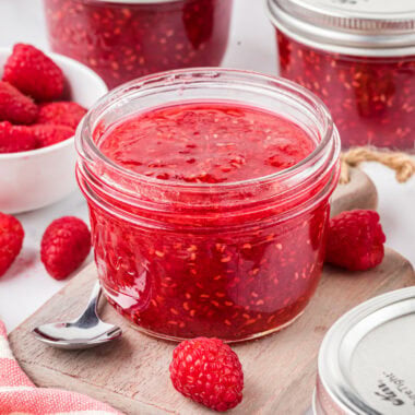 In the center of the image is an opened jar of jam ready to serve with other unopened jars and raspberries in the foreground and background.