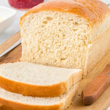 In this image, the bread is on the cutting board, with two slices cut in front of the loaf. It has butter and jam in the background.