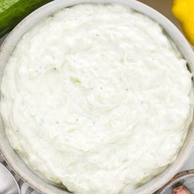 In this overhead image, the tzatziki sauce is in a serving bowl ready to serve with additional cucumber, lemon, and garlic surrounding it.