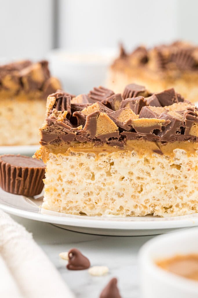 In this image, the bars are cut and plated. The bar in the front is the central focus. In the forefront of the image are a few chocolate chips, rice krispies, and a small bowl of peanut butter. In the background, there are two other plated treats.