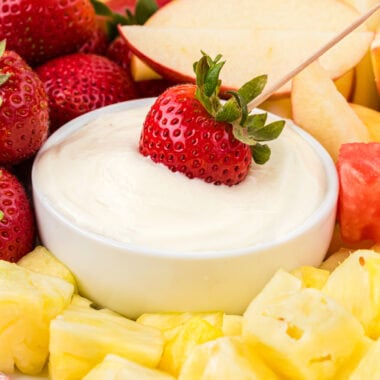 In this image the dip is in a serving bowl on a platter with strawberries, apples, pineapple, and watermelon around it. a strawberry with a toothpick is placed in the center of the dip ready to eat.