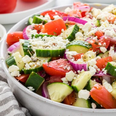 The salad is dressed and ready to serve in a bowl in the center of the frame with additional tomatoes, red onion, and cucumbers in the background.