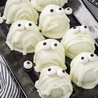 In this image, the mummies are arranged on a rectangular platter, with the mummies looking up. Extra Oreo balls and candy eyeballs are in the background. A black and white striped linen wraps around the bottom of the platter.