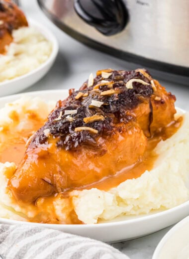 In this image, the cranberry chicken is set over a bed of mashed potatoes on a plate in the center of the image with a crockpot and other plate in the background.